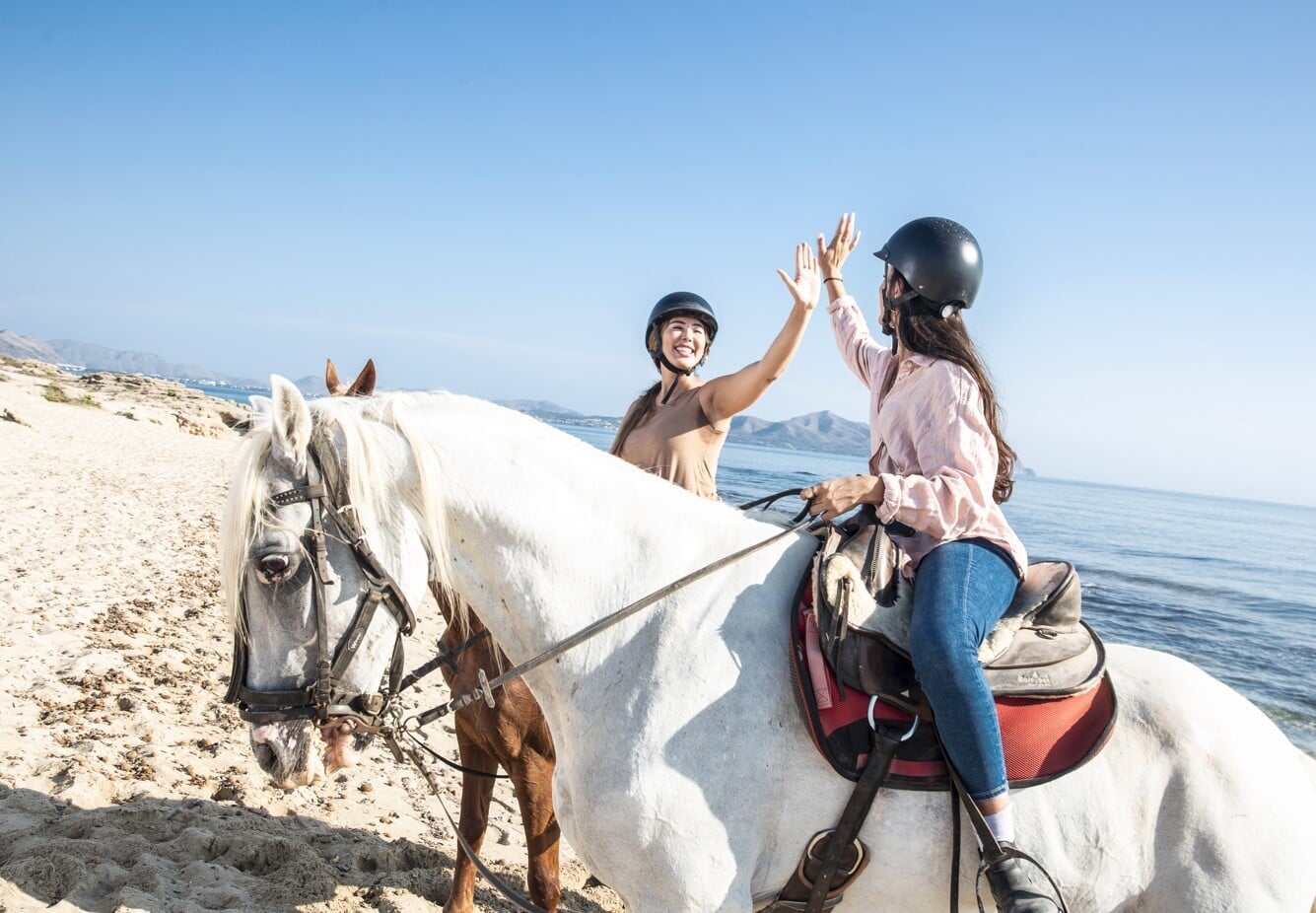 Amigas montando a caballo en la playa en un día soleado en Mallorca celebrando juntas junto al mar