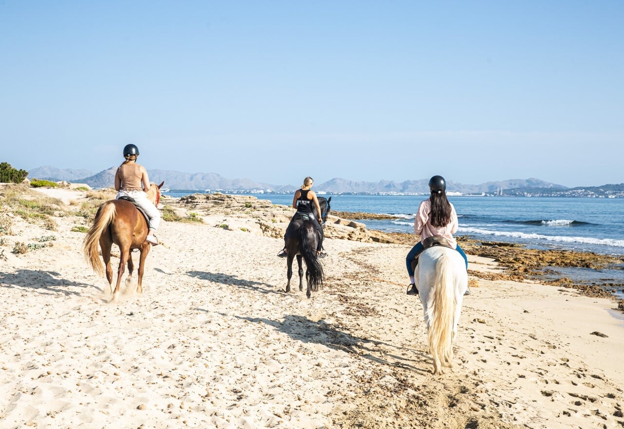 Tres personas cabalgando en la playa junto al mar con caballos negro, marrón y blanco durante una excursión a caballo